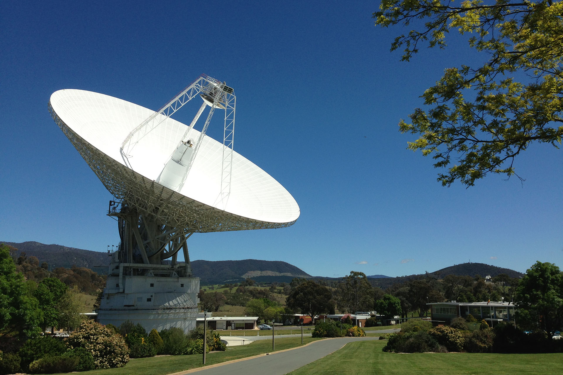 Deep Space Station 43, a massive white antenna, points to a clear, bright blue sky. Mountains stretch across the horizon in the background.