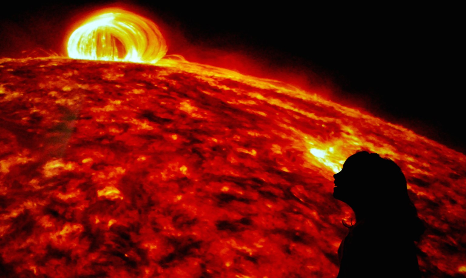 An excitedly smiling child appears silhouetted in profile against the backdrop of an image of the Sun, its surface curving across the screen from left to right and a bright yellow loop erupting from its red and yellow swirling surface. Space is black behind the Sun, and the Sun's corona appears as a diffuse red glow along its edge.