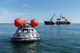 An Orion capsule, with 3 red inflated buoys on top, floats in the ocean near a large Coast Guard ship