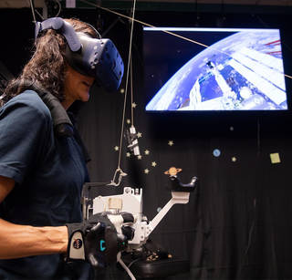 Astronaut Sunita Williams wears virtual reality headgear and gloves while using a simulator while a screen with the ISS