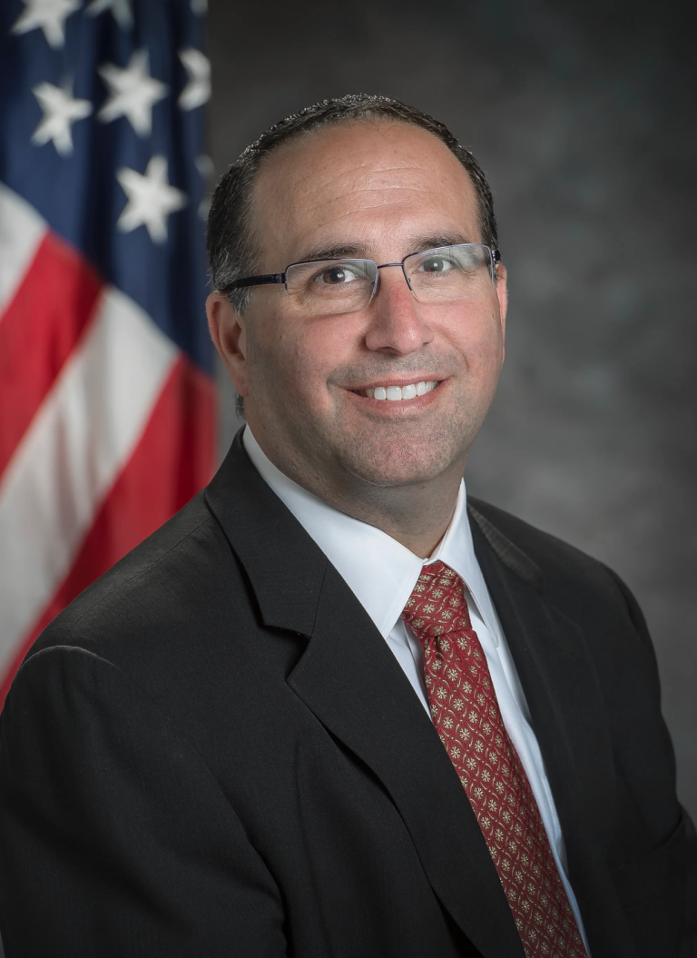 Dr. Erik Weiser, Deputy Assistant Administrator of Office of Strategic Infrastructure, poses in front of American flag wearing suit and tie. 