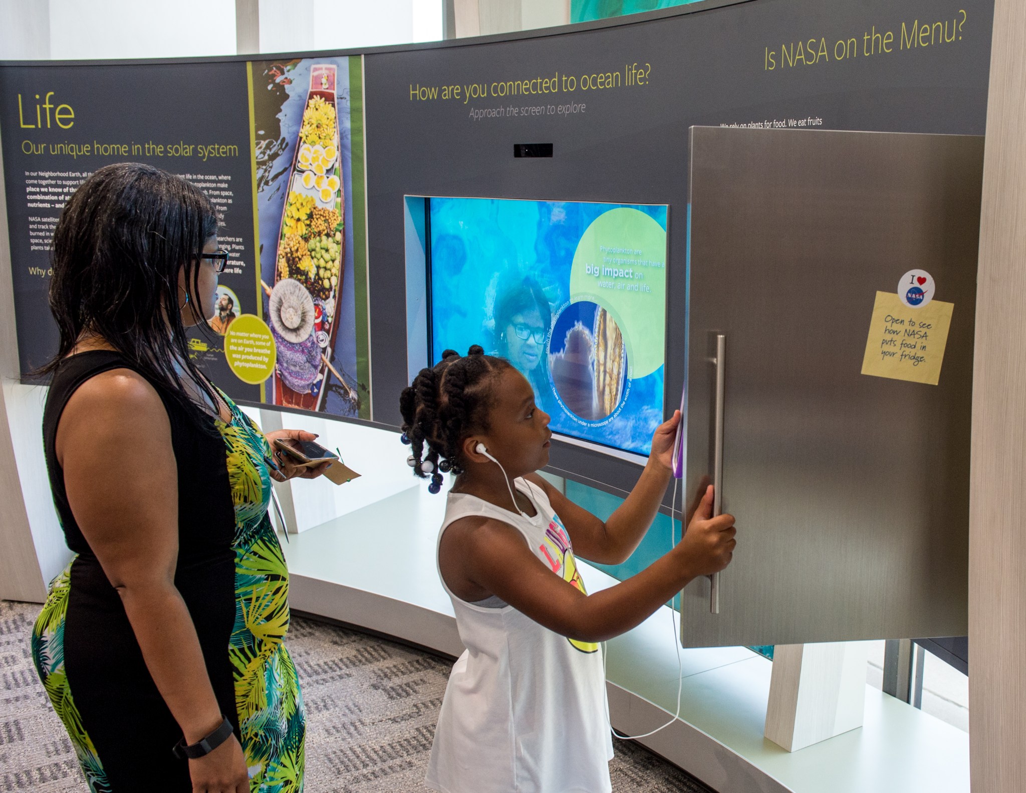A young Black girl wearing a white tank top opens a silver door in a wall-mounted museum exhibit, holding a purple phone with white earbuds plugged into her ears. The exhibit discusses ocean life on Earth, and the silver door opens like a refrigerator to an exhibit about food. A Black woman wearing a black and green dress looks on.