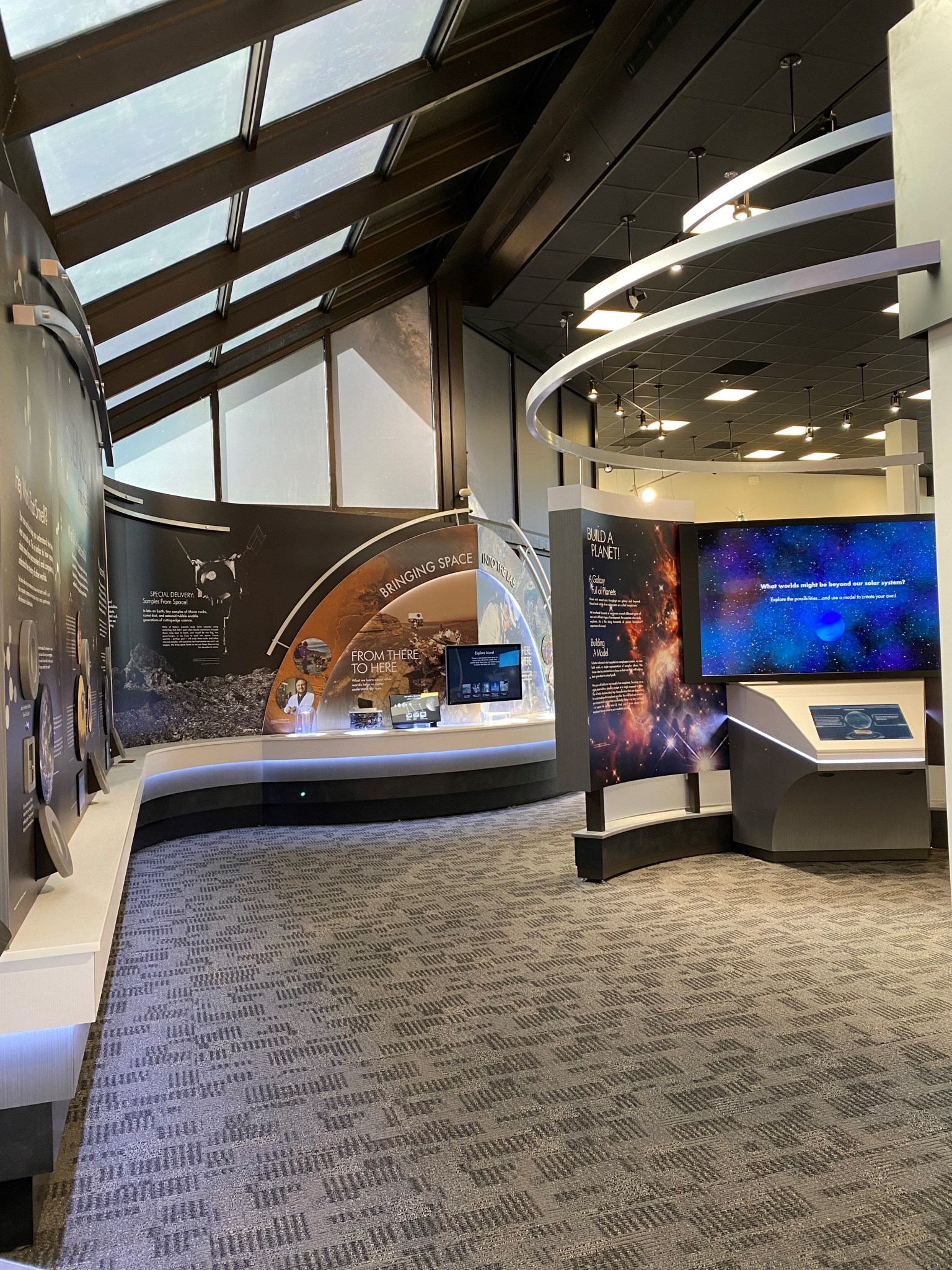 Wall panels from a colorful space museum exhibit line the walls of a large room with tall windows and skylights, a black ceiling, and gray carpet. The panels feature colorful photos of galaxies, nebulas, and spacecraft landing on the Moon and Mars.