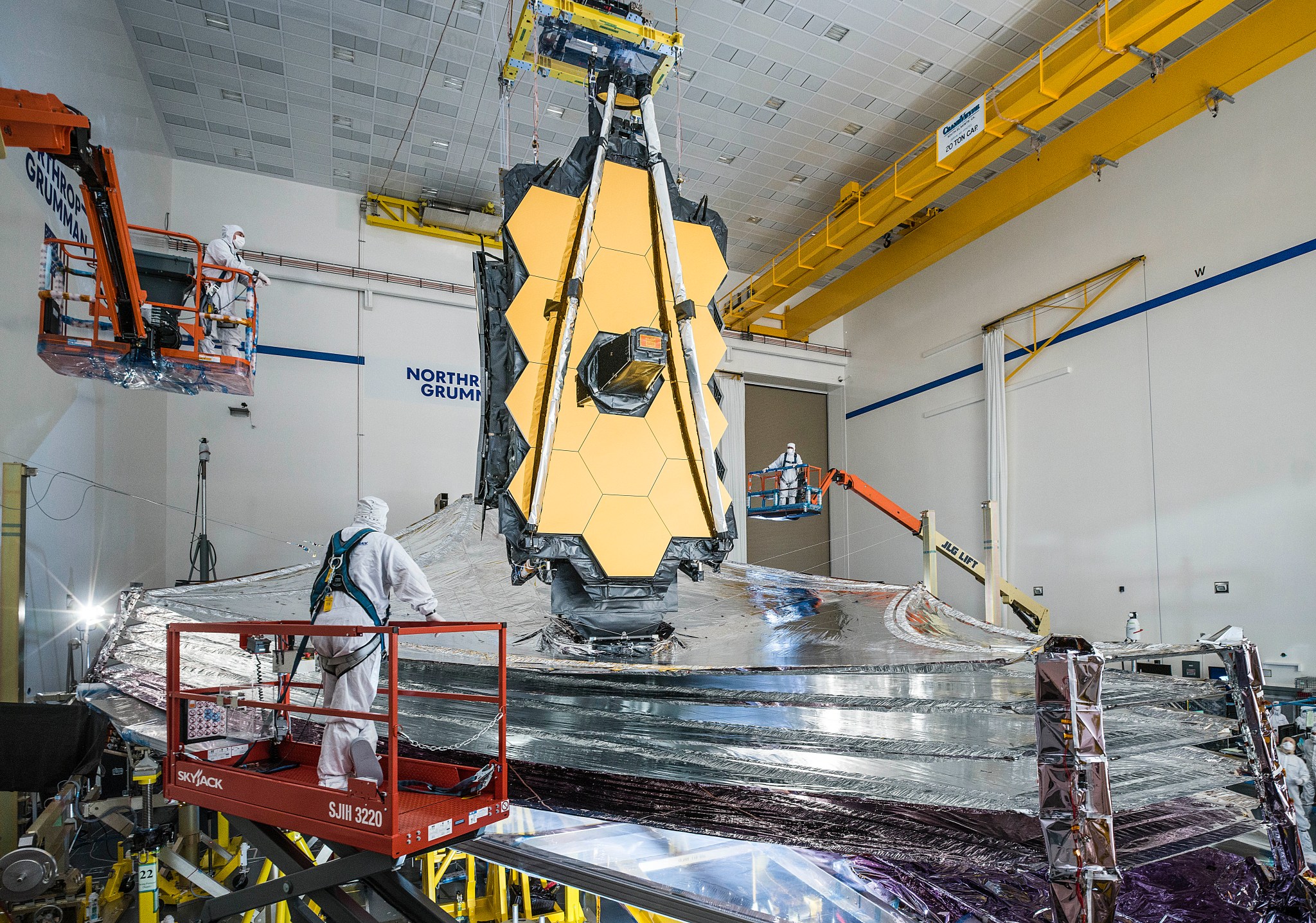 The James Webb Space Telescope, comprised of a flat, multi-layered silver sunshield stretched out horizontally like sails underneath a vertical strip of gold hexagonal mirror panels. The telescope is under construction, with two people in white coveralls standing on red platforms overlooking the white clean room where the telescope is sitting. Blue text reading "Northrop Grumman" is partly visible on the back wall.