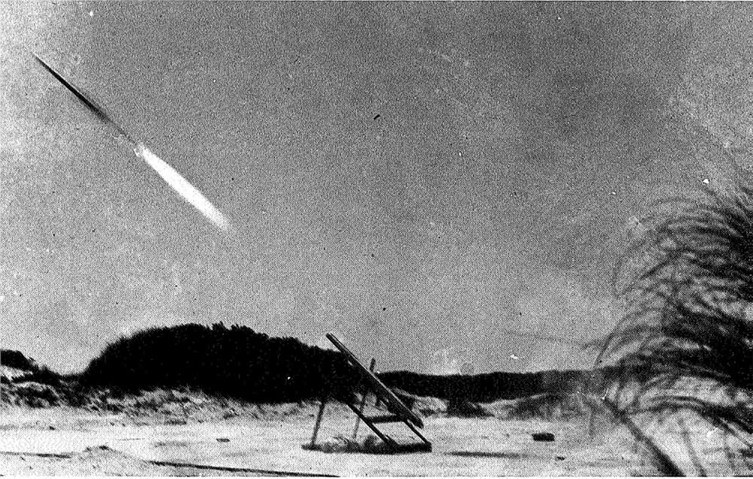 A black and white image of a small rocket taking off from a stand on a beach. The rocket is blurry, launching into the left side of the image with a small white flame behind it. A small stand is in the center of the image, with some vegetation behind it.