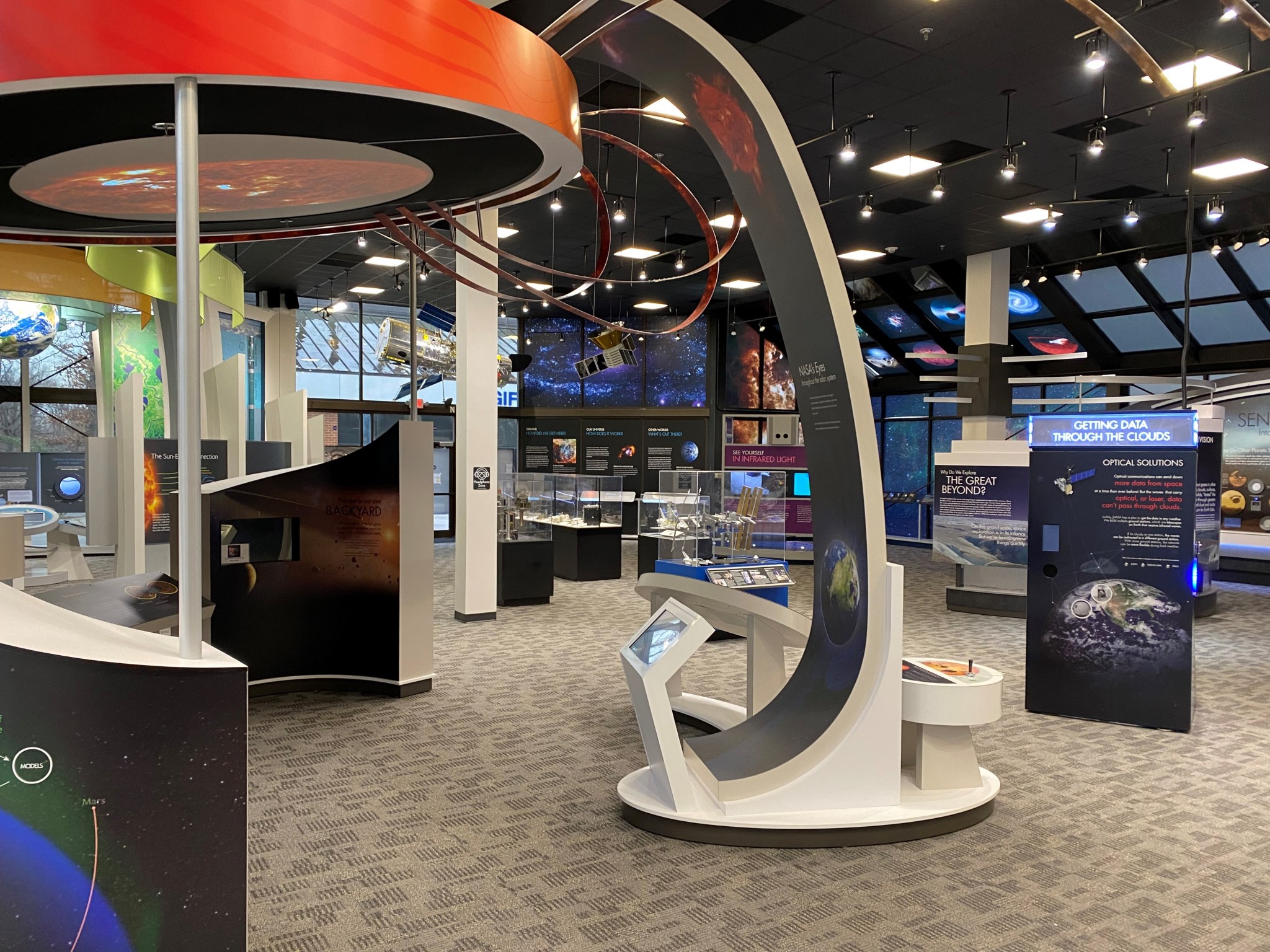 A museum exhibit with colorful signs and displays in shades of blue, purple, and black. In the foreground, a stylized model of the solar system features a flat, red circle mounted horizontally near the ceiling, with graceful arched supports containing text about the Earth and Sun, and shiny red loops curving out from the Sun's red circle.