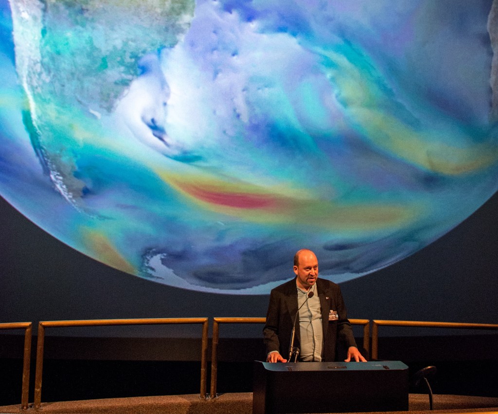 GISS Director Gavin Schmidt stands at a podium, giving a talk. He is dwarfed by a massive screen behind him, projecting a huge globe Earth. Earth has data visualized on it to show wind currents in the atmosphere. Visible from this view are South America and part of Antarctica and the Atlantic Ocean, with streaks of red, yellow, green, and blue indicating different datasets.