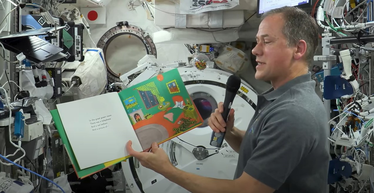 NASA astronaut Thomas Marshburn, holding the book 'Goodnight Moon' in his left hand and a microphone in his right hand, reads aloud aboard the International Space Station.