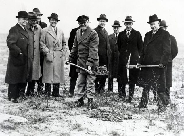Black and white photo of a group of men wearing fedoras and overcoats. Two men in front hold shovels filled with dirt.