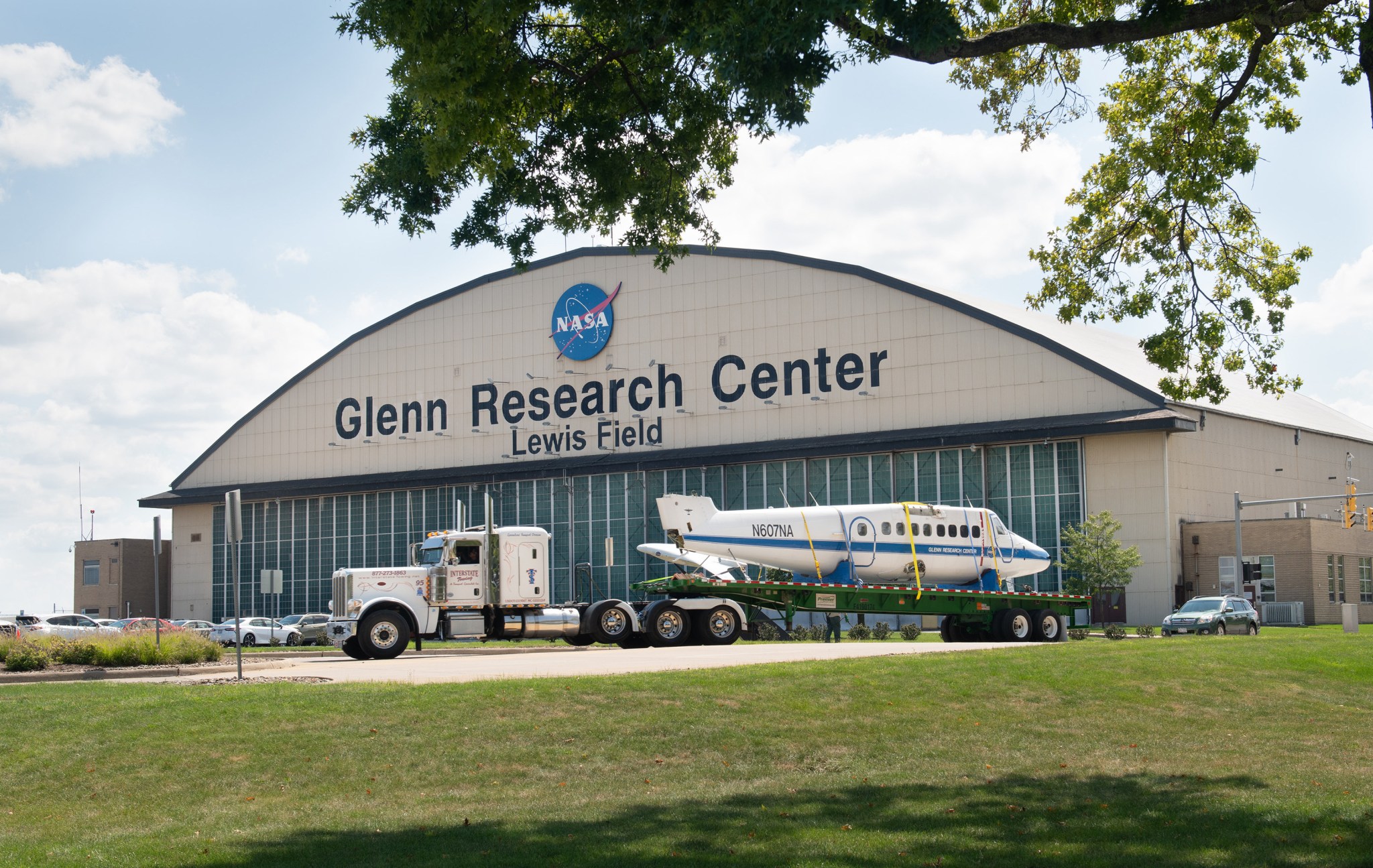 Body of aircraft sits on a flatbed attached to a truck in front of the NASA Glenn Research Center aircraft hangar.