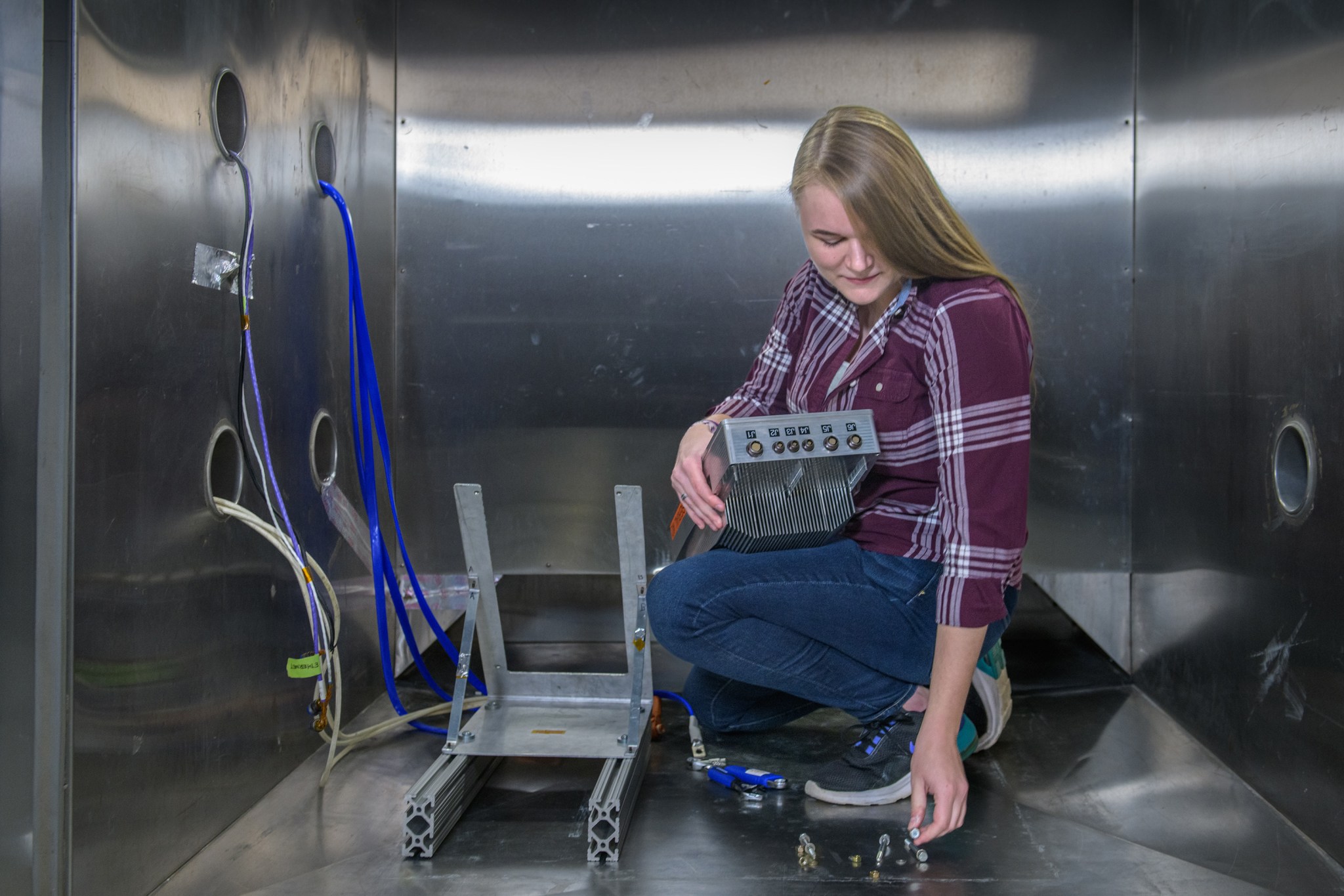 Woman in flannel shirt, jeans and sneakers bends down to pick up bolts next to testbed architecture inside metal wind tunnel.