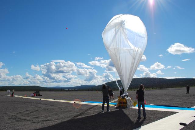 People stand around a partially inflated scientific balloon, which is plastic, see-through in the shape of an upside down teardrop. There is a small mountain range in the background and bright blue sky with a few clouds.
