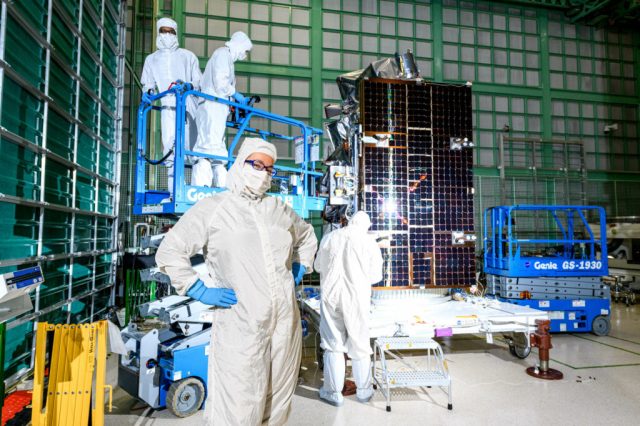 A woman stands in the center left of the image wearing a white clean suit which covers her whole body including her head. She wears a white mask that covers her nose and mouth and blue gloves on her hands. Behind her are three other people wearing the same clean room gear. Two of those people are standing on a lift and are slightly above the woman in the foreground. Behind all of the people is PACE, which is large and box shaped, and is black colored with a light reflection off of it.