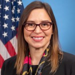 Portrait of Makenzie Lystrup, smiling at the camera. She has medium brown hair, lighter closer to the ends. She is wearing brownish, plastic-rimmed glasses, and a black, red and gold scarf, and black blazer. Behind her is the American flag.