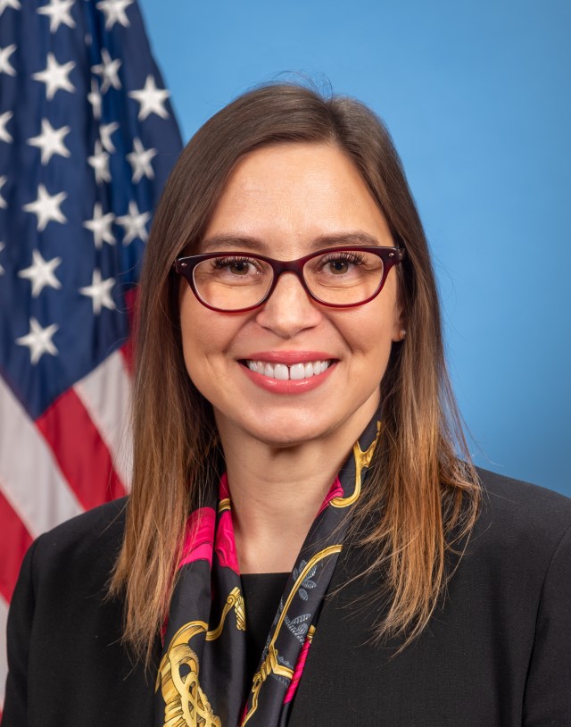 Portrait of Makenzie Lystrup, smiling at the camera. She has medium brown hair, lighter closer to the ends. She is wearing brownish, plastic-rimmed glasses, and a black, red and gold scarf, and black blazer. Behind her is the American flag.