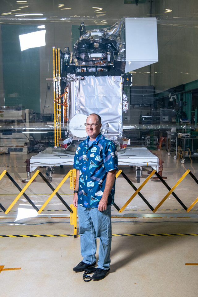 A man stands centered in the image wearing jeans and a Hawaiian-like blue button-up shirt. Behind him is a clear, plastic screen separating him from the room behind him. Past the screen, PACE sits. It's bottom half is covered in a silver-colored foil-like material.