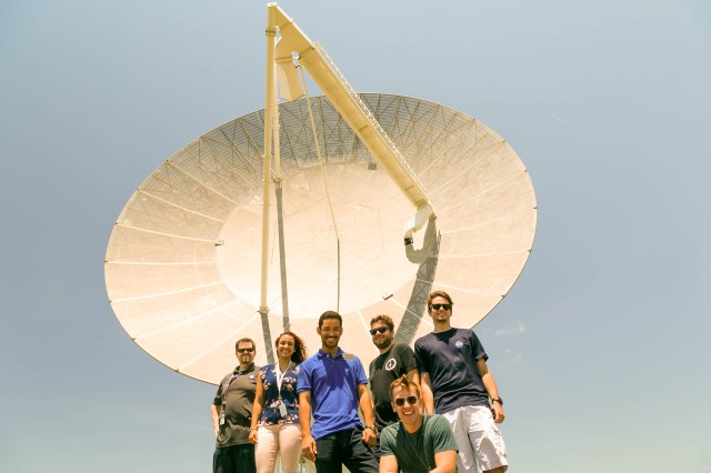 A group of interns stand beneath an antenna and a bright blue sky.