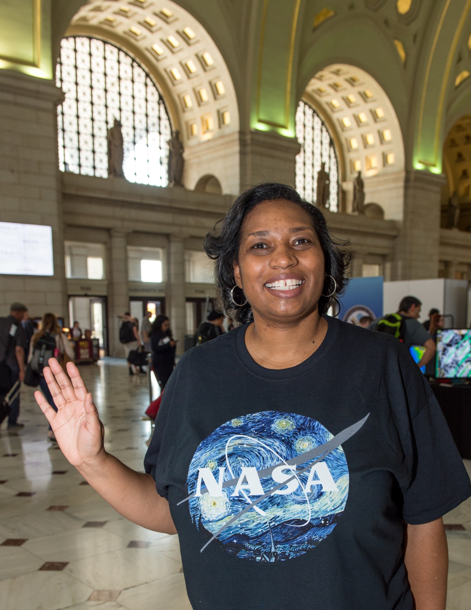 Trena Ferrell smiles and waves at the camera in a large train station atrium with high, arched ceilings. She wears a navy tee with a colorful NASA logo and hoop earrings. Many people are visible in the background.