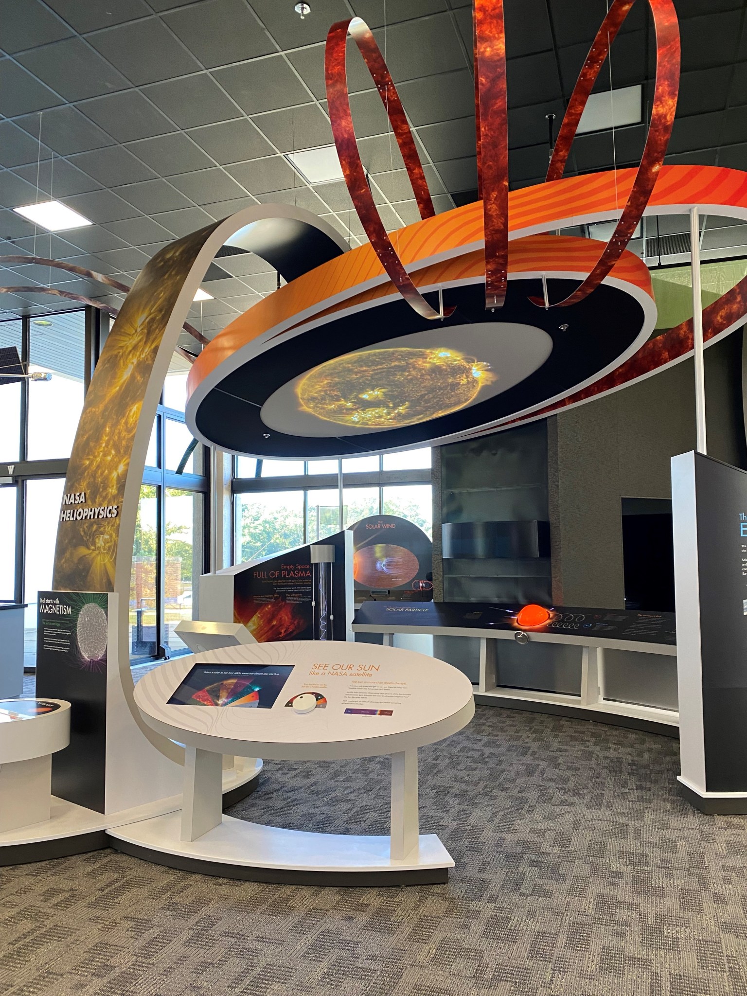 A colorful museum exhibit depicting the Sun as a large, horizontal circle with orange "orbits" and loops surrounding it, mounted on a gracefully curved white pillar. Large windows with visible sky and trees are in the background.