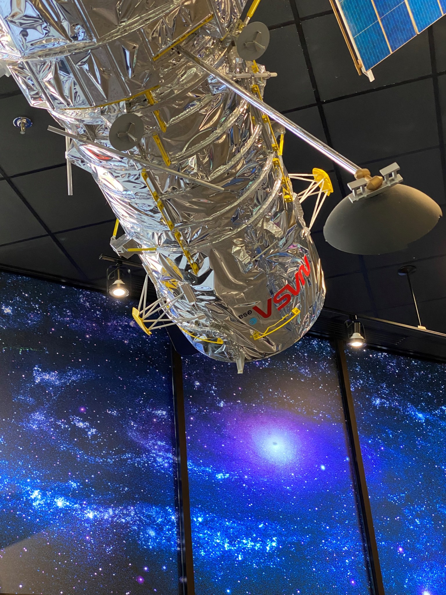 A model of the Hubble Space Telescope, a silver cylinder with an instrument arm sticking out from its center, hangs from a black ceiling in front of a lit mural of blue, purple and white galaxies.