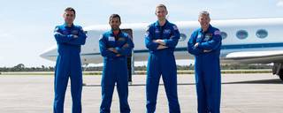 Four male astronauts pose with arms crossed in front of a plane in blue flight suits on the tarmac.