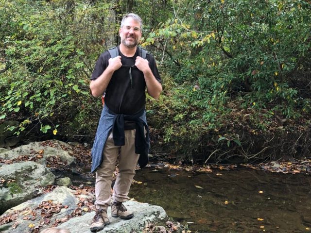 A man stands in the left of the image wearing light brown pants and a black T-shirt. He stands on a rock next to a small body of water. In the background there are green leaves and foliage. The man stands looking at the camera.