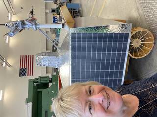 A selfie of a woman with short blond hair in front of a model of a Moon rover and an American flag behind. The rover is silver in color with solar panels on its side and gold-colored wheels. The mast on top is taller than the woman.