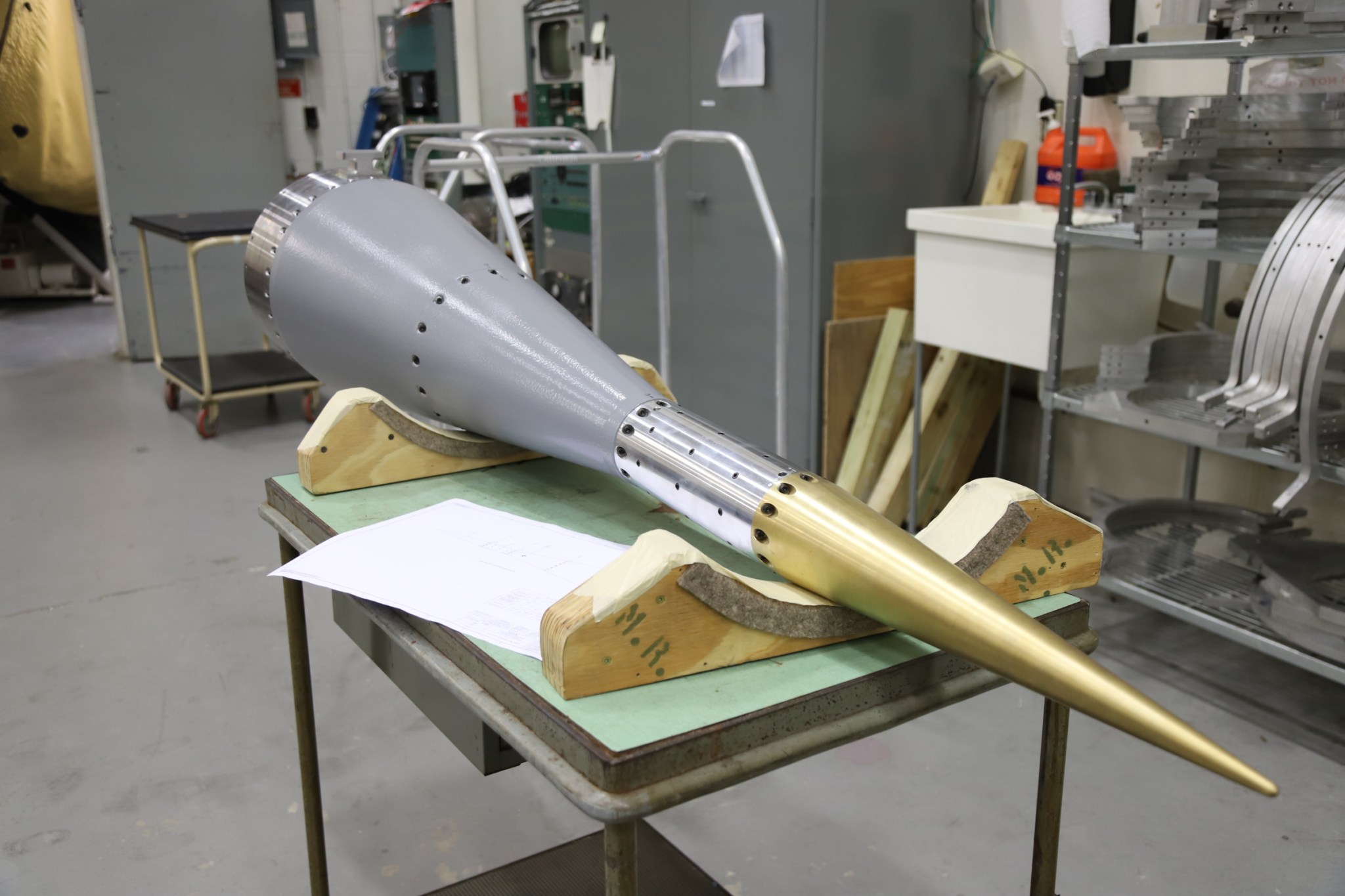 The payload section of a sounding rocket propped up on a table. The payload is mostly grey on the larger section and gets small to a golden pointed end. 