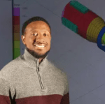 Man in sweater stands in front of 3D technical drawings while looking at the camera.
