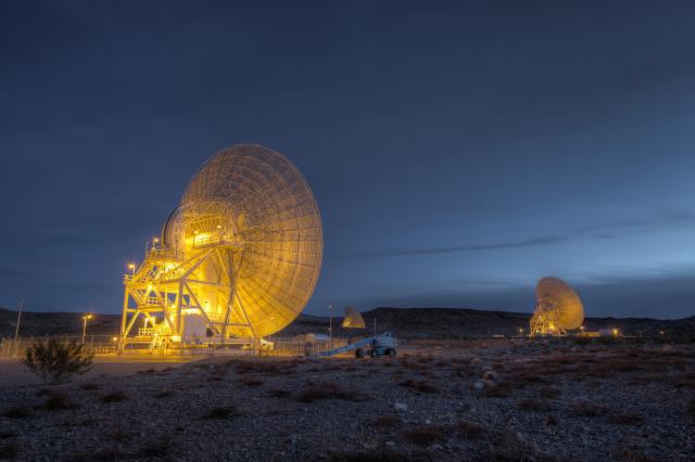 Three Deep Space Network antennas light up the night sky with golden hues. The antennas face away from the viewer.