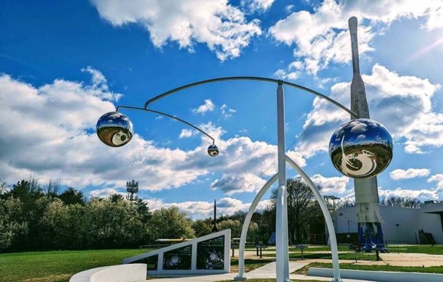 A colorful photo of a metal sculpture, with three shiny silver globes hanging from gracefully balanced, curved supports. One large silver globe hangs on the right side of the center support, and the left side supports a smaller bar holding two smaller silver globes. The three represent the Sun, Earth, and Earth's Moon. The sculpture stands outdoors in a cement circle surrounded by printed placards with information about Earth and weather observations. A tall, old-looking rocket stands in the background. The sky is deep blue with white clouds, and grass and trees are visible further back.