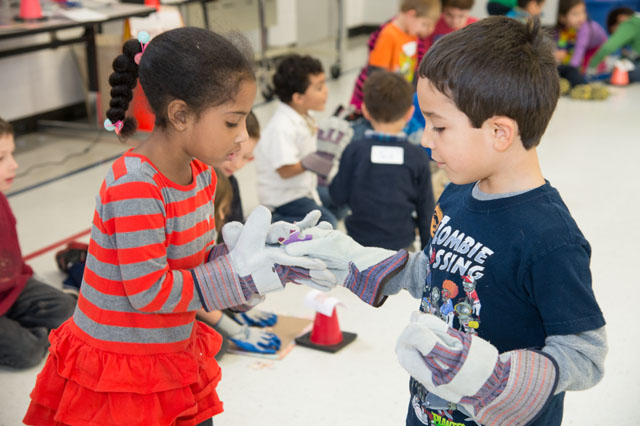 Two kids are wearing thick gardening gloves. One is holding and looking at one of the gloves of the other.