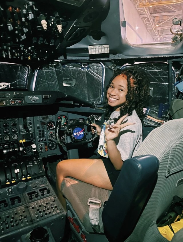 A teenager sits in the cockpit of a craft, and is flashing a peace sign at the camera.