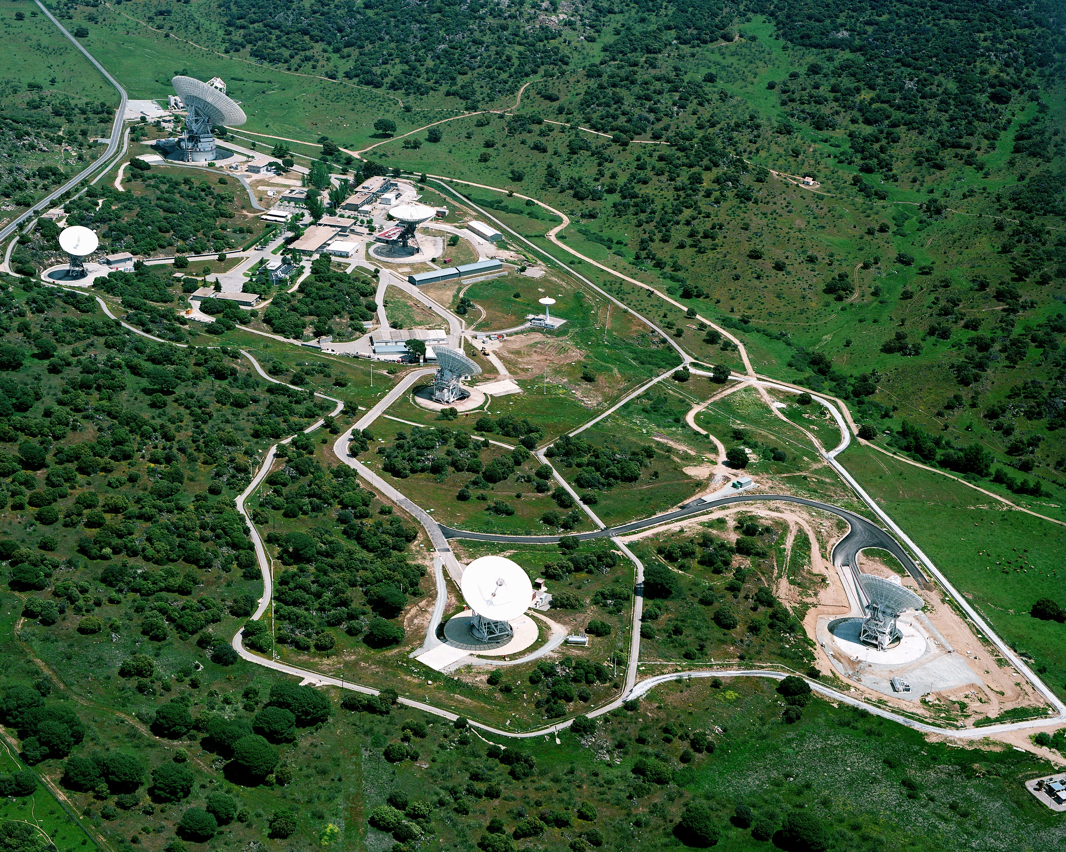 The Madrid Deep Space Communications Complex stretches across a lush, green landscape. Several antennas can be seen towering over surrounding buildings and roads.