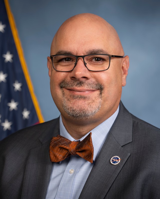 Joseph Pelfrey, a man with glasses and a bow tie stands in front of the American Flag