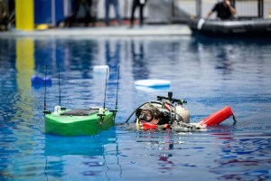 Diver in the Neutral Buoyancy Lab floats above the water, pushing a green, floating apparatus