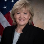 A photo of Jody Singer from Marshall Space Flight Center in front of an American Flag wtih a brown background behind her.