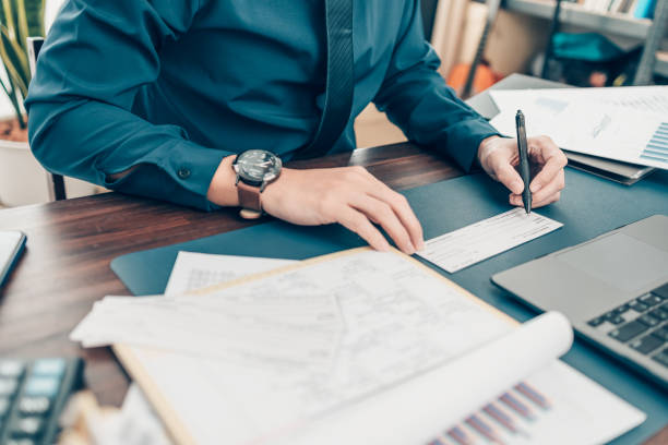 A person holding a pen works at a desk filled with papers. The corner of a laptop can be seen in the righthand corner of the image