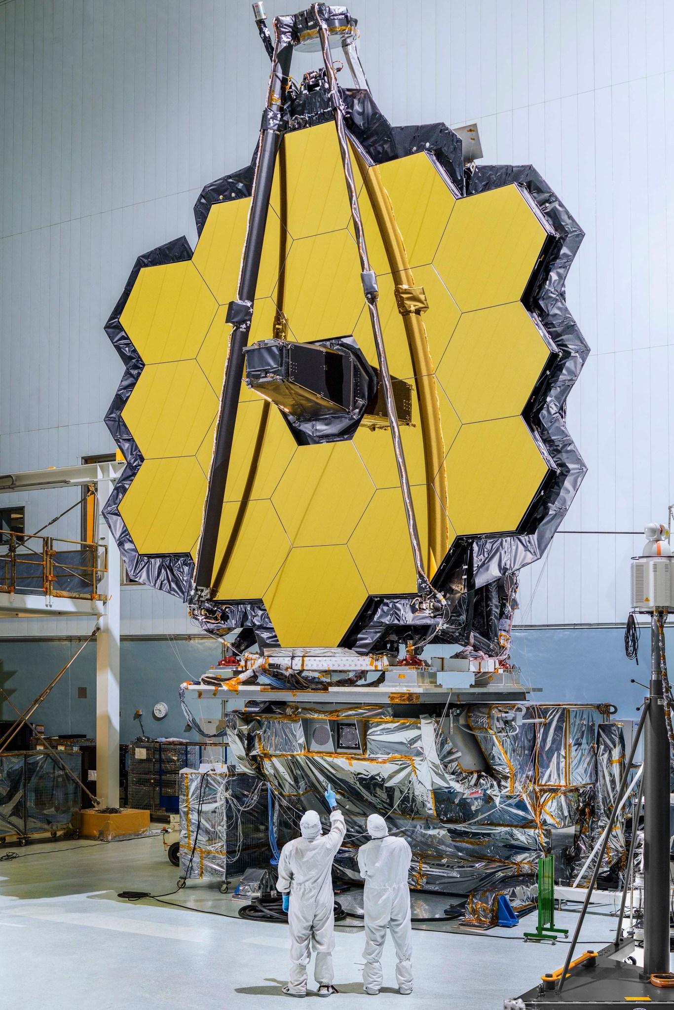 Two people in white coveralls look up at the large primary mirror of the James Webb Space Telescope. The mirror is made up 18 gold-coated mirror segments surrounding a black nose cone. The long silver "arms" of its secondary mirror support structure are folded upwards, forming an upside down V on top of the primary mirror. The mirror stands in a white clean room atop a mount covered with shiny, crumpled protective blanketing.