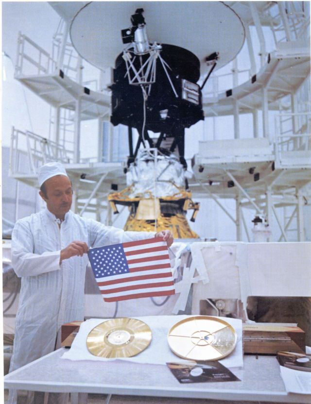 Man in clean room holds American Flag over Voyager Golden Record. 