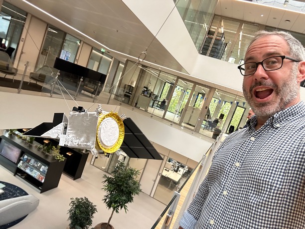 A man wearing glasses and in a light blue checker patterned button up shirt takes up the right side of the image. He is taking a selfie and smiling at the camera. The left side of the picture is aimed over the edge of a balcony inside of a building. Hanging from the ceiling is a model of a spacecraft - PACE. Behind the spacecraft, parts of the floor below, the floor the man is on, and the floor above can be seen.