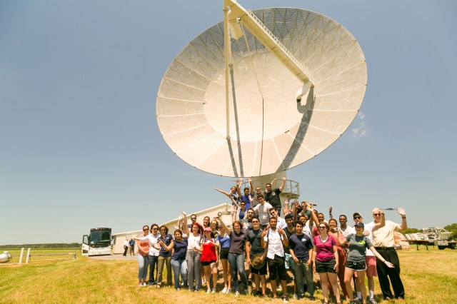 A large group of interns stand beneath an antenna and a bright blue sky.