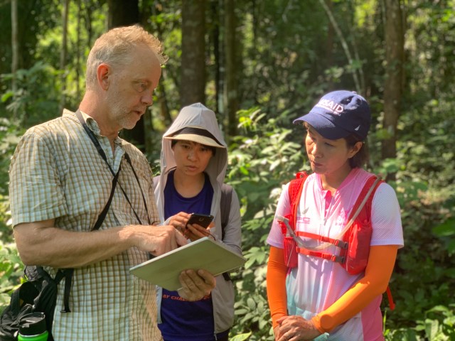 A man holds an iPad and is showing two women in hiking hear the screen.