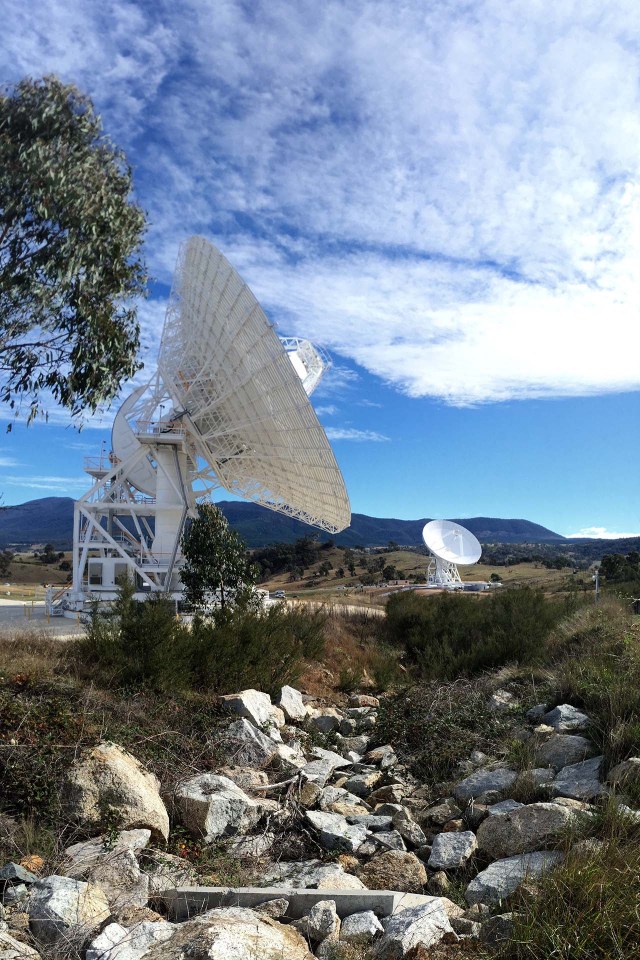 Two antennas point to a bright blue sky covered with fluffy white clouds. A mountain range stretches in the background. In the foreground, a landscape of rocks and greenery extend toward the viewer.