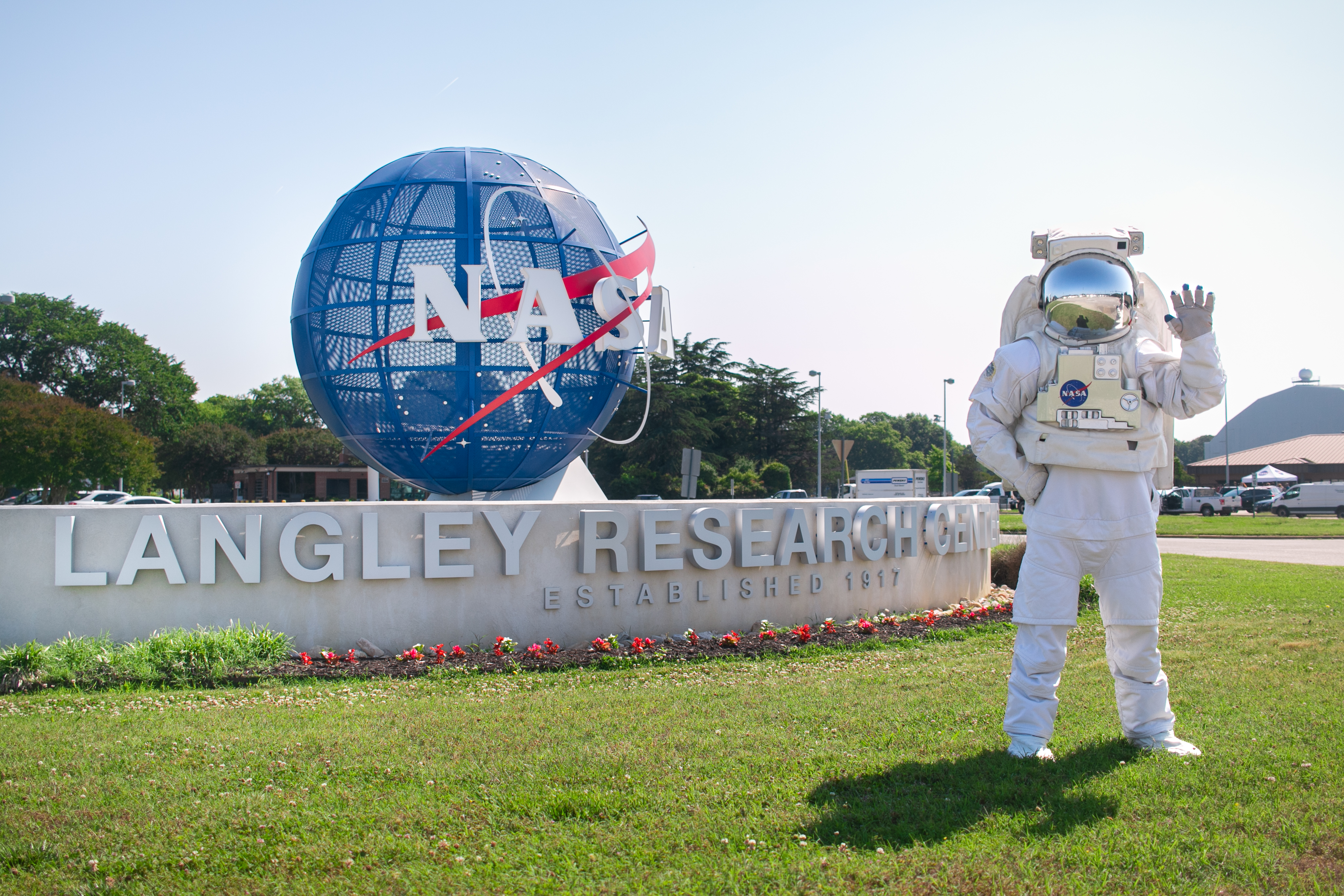 A NASA employee wears an astronaut suit and stands on bright green and freshly cut grass in front of the welcome sign at NASA Langley Research Center. The welcome sign features a large, blue globe with the NASA insiginia on it and text on a long stone ridge that reads "Langley Research Center." Flowers line the edge of the stone ridge.