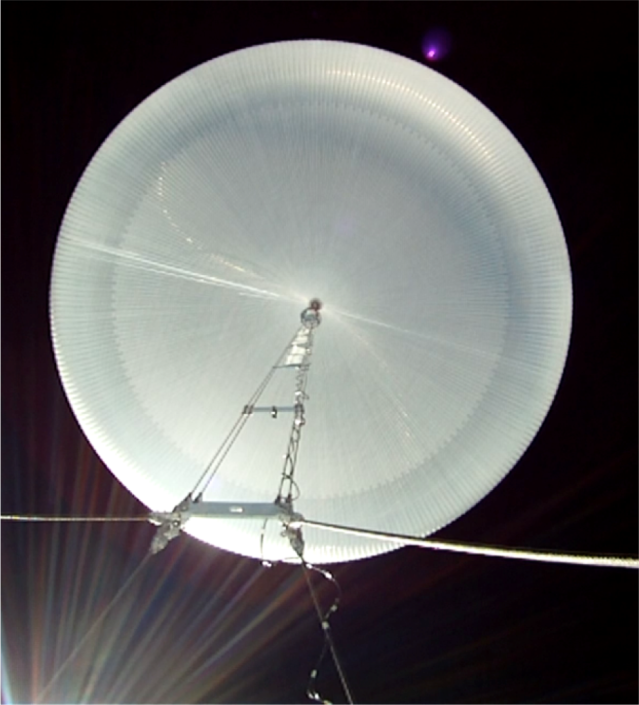 An image of a fully inflated scientific balloon floating in the atmosphere. The image is looking up from below the balloon. The balloon is a clear, plastic perfect circle.