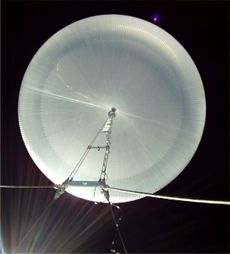 An image of a fully inflated scientific balloon floating in the atmosphere. The image is looking up from below the balloon. The balloon is a clear, plastic perfect circle.