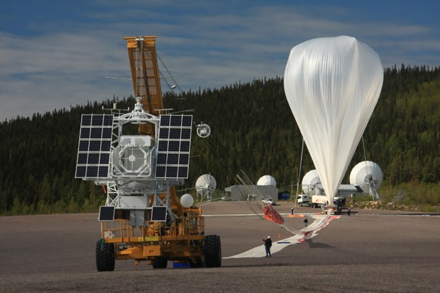 A crane is leading a scientific balloon before launch. The balloon is to the right, and appears an a plastic, upside down teardrop. A tube attached to the top of the balloon leads down to the ground where a number of personnel are holding it. To the left, a crane holding a large payload structure with many solar panels is attached to the end of the balloon.