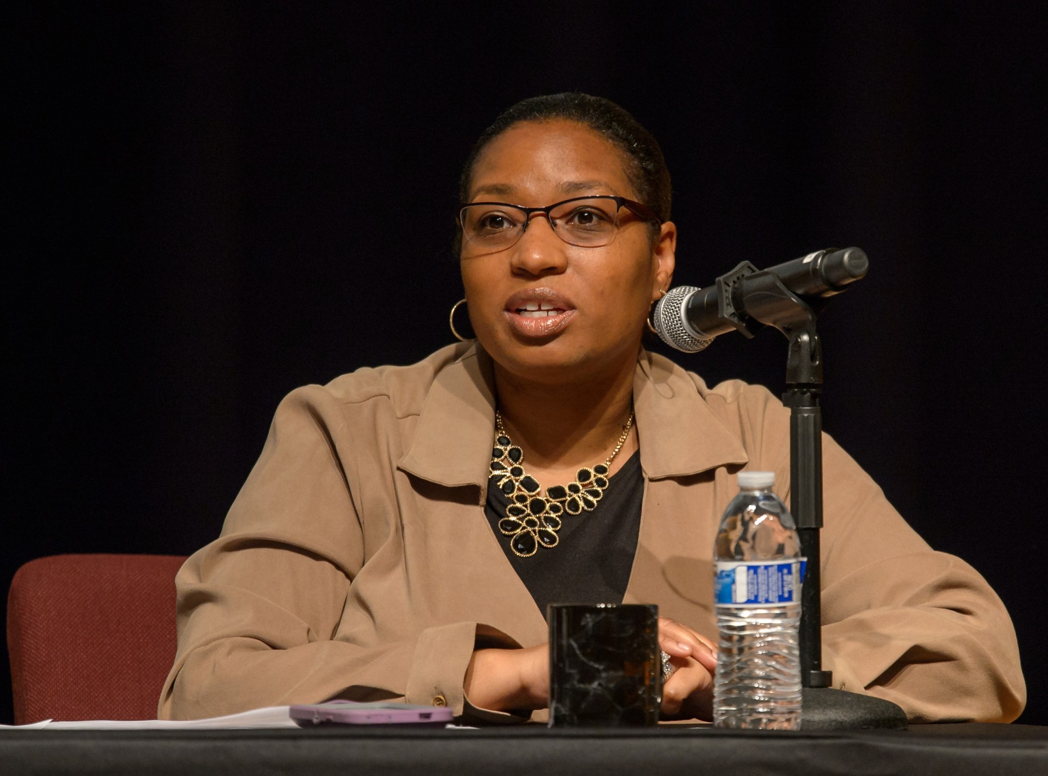 Trena Ferrell sits at a table and speaks into a microphone, looking slightly left and off camera. She wears glasses, hoop earrings, a black-and-gold necklace, and a tan jacket with black tee.