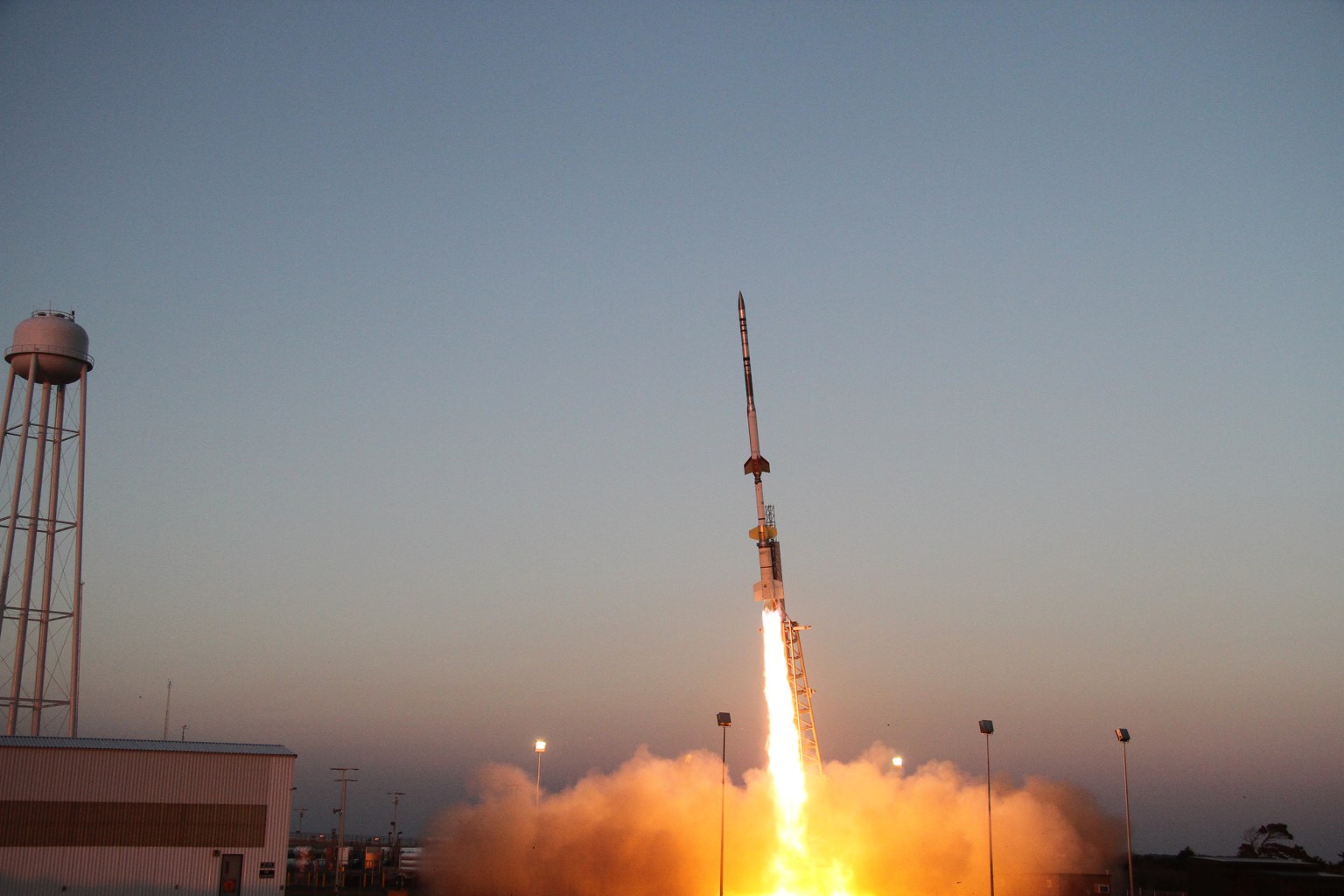 A sounding rocket seconds in mid-launch off the launch pad with a bright yellow plume underneath against a pale blue sky.